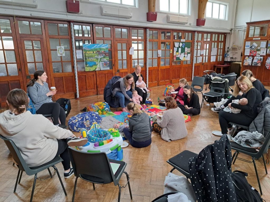Parents and babies sitting in a circle on the floor of a wood-panelled community hall at a Breastfeeding Together group