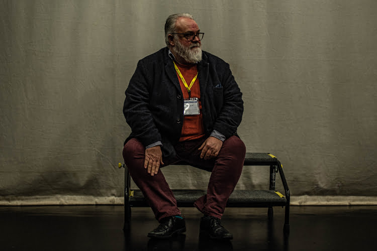 A Made in Wigan portrait: a bearded man in a dark jacket and lanyard seated on a bench against a plain backdrop