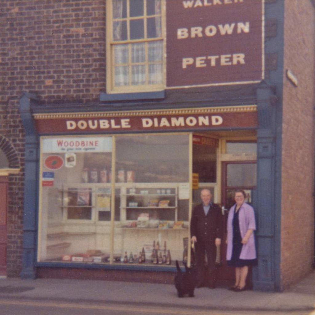 An archive photograph of an Abram Ward corner shop with a Double Diamond sign, an elderly couple and a black dog standing outside