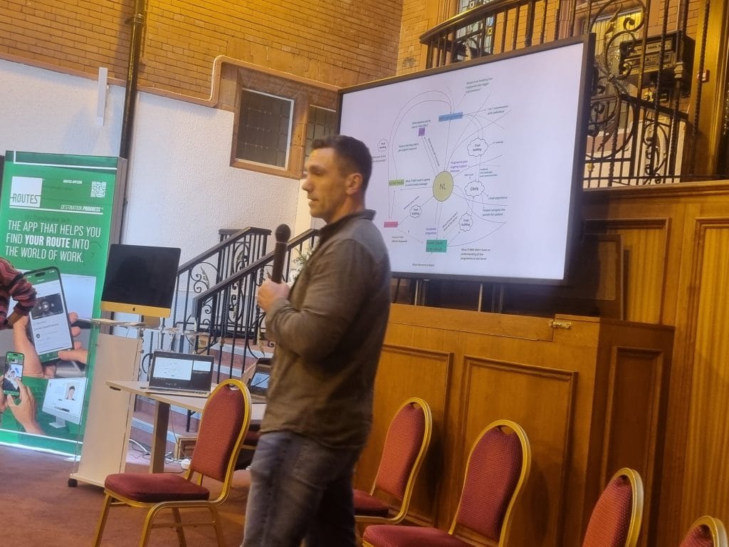 A speaker presenting a diagram on screen at Social Enterprise Day, beside a banner for the Routes careers app