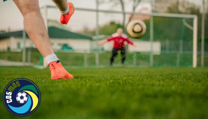 A footballer striking the ball towards a goalkeeper on a grass pitch, with the Wigan Cosmos club badge in the corner