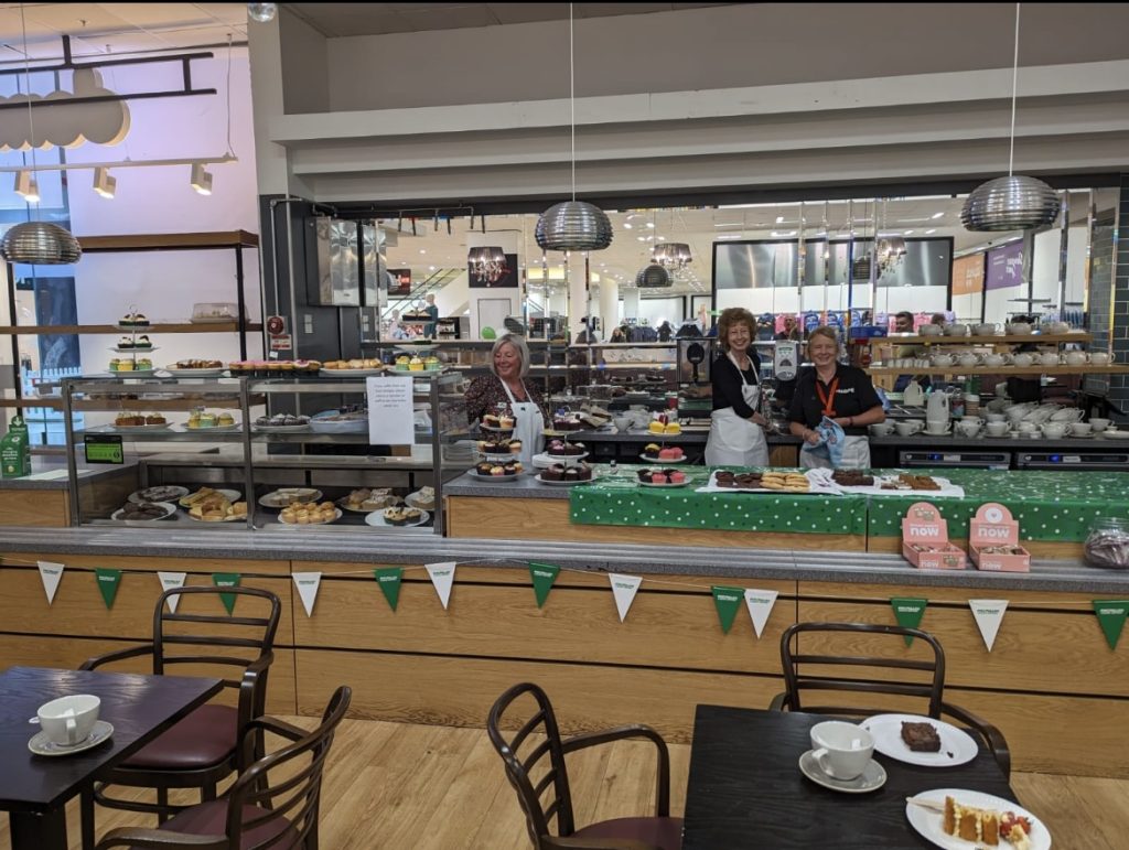 Staff behind the counter of the café, with cakes and pastries on display and bunting overhead