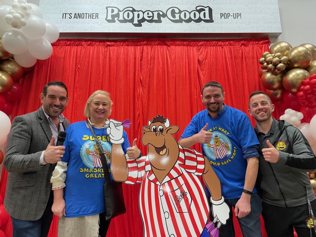 Four guests giving a thumbs up beside a cartoon cut-out at the Proper Good pop-up launch, against a red curtain and balloon arch