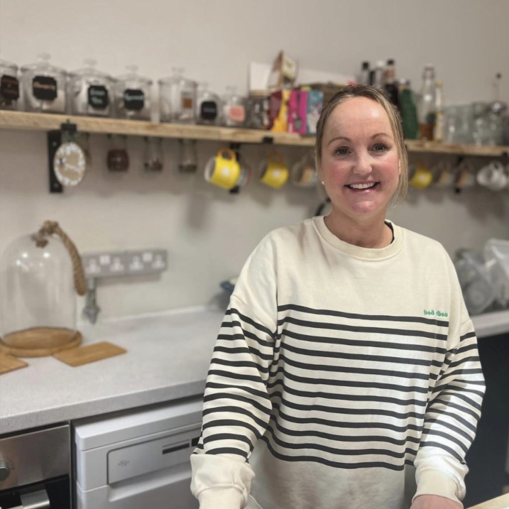 A smiling team member in a community kitchen, with storage jars and mugs on the shelves behind