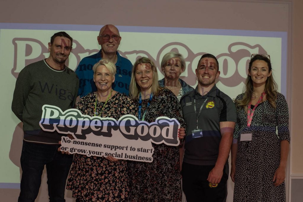 A group of partners holding the Proper Good sign in front of a Proper Good presentation screen