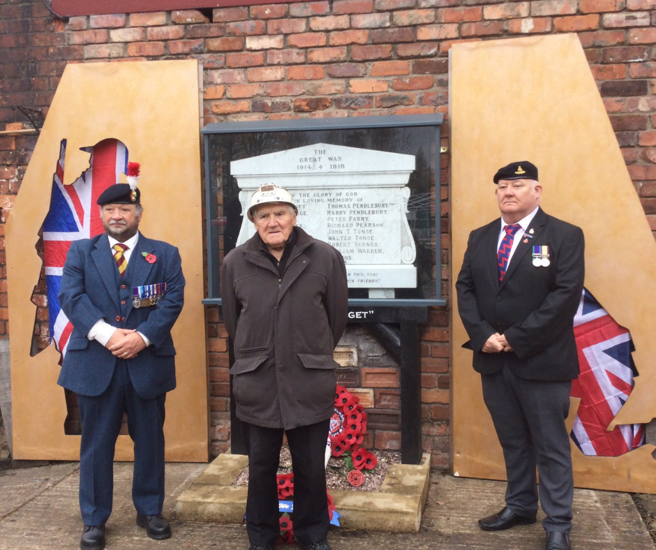 Two uniformed veterans and a resident standing either side of a restored Great War memorial, with poppy wreaths laid beneath